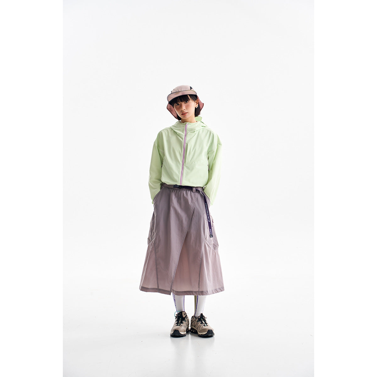 Model wearing a pale green hooded jacket and a mauve wrap skirt with sneakers and a bucket hat.