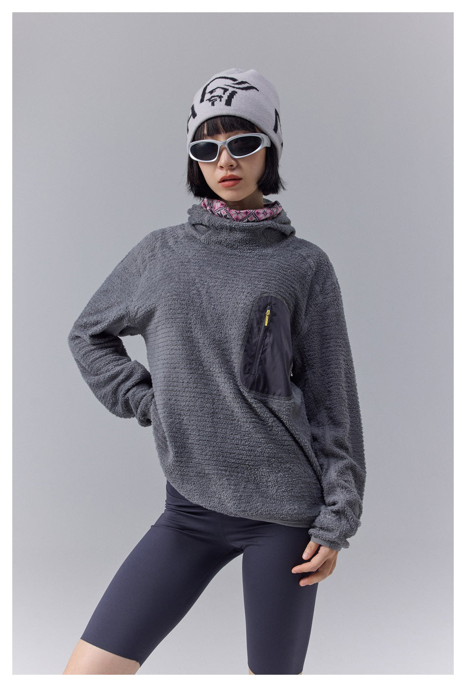 Model wearing a charcoal gray textured hooded pullover with a dark zip chest pocket and navy cycling shorts.