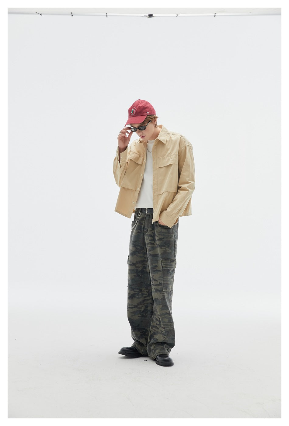Model in a beige button-up overshirt and loose dark camouflage cargo pants, adjusting his sunglasses.