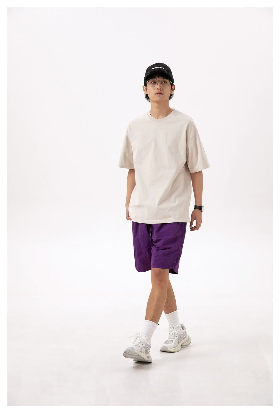 Man walking in a sand beige oversized crew neck T-shirt with purple shorts, cap and white sneakers