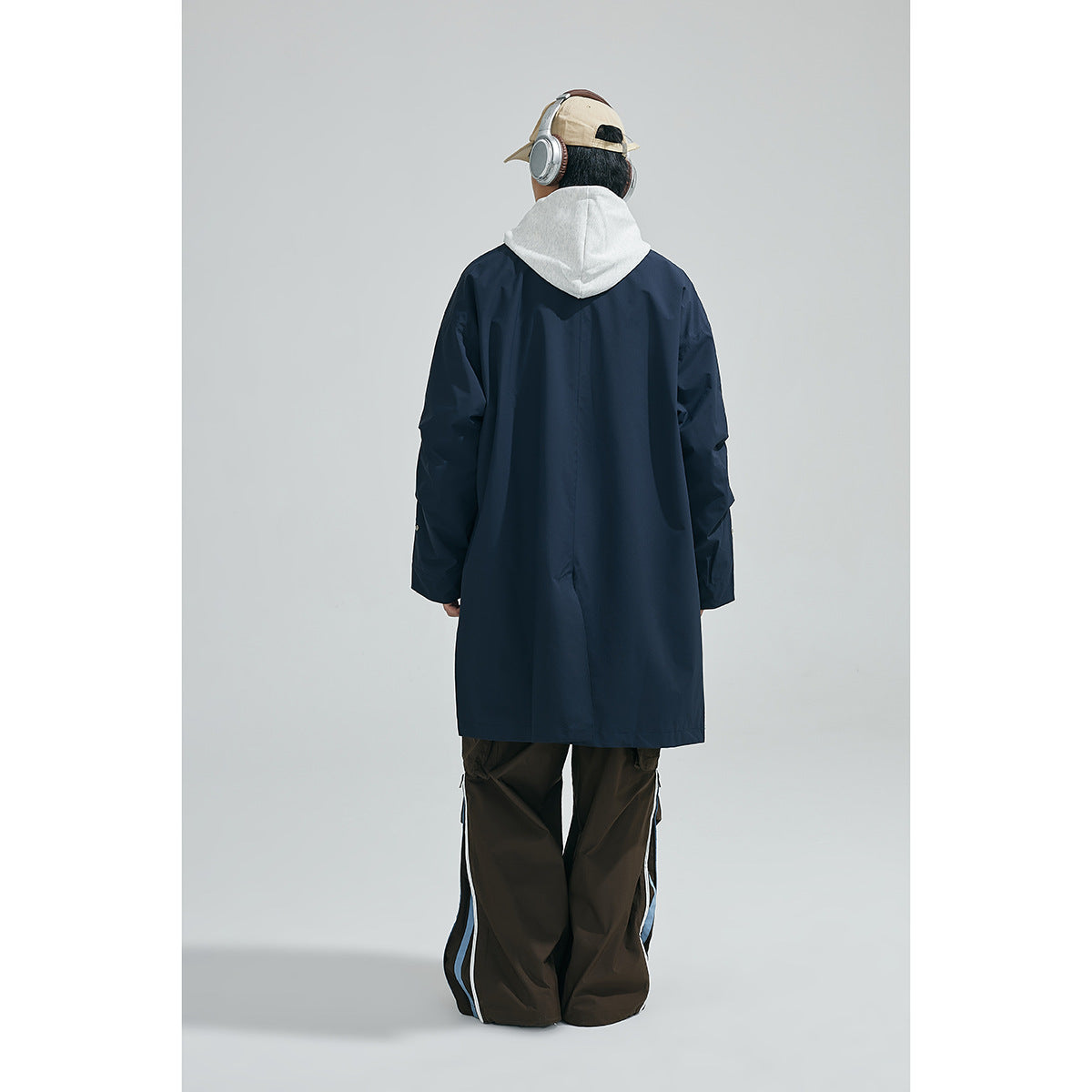 Rear view of a model wearing a long navy coat over a light gray hood, with dark trousers.