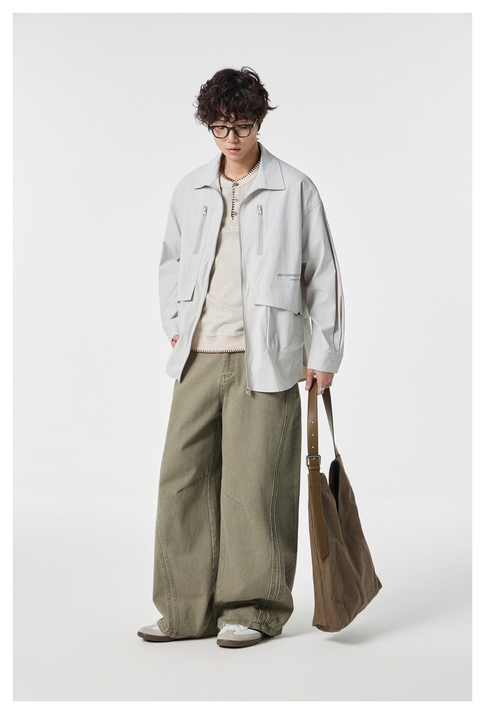 Model wearing an open pale gray utility jacket and loose olive trousers, carrying a brown shoulder bag.
