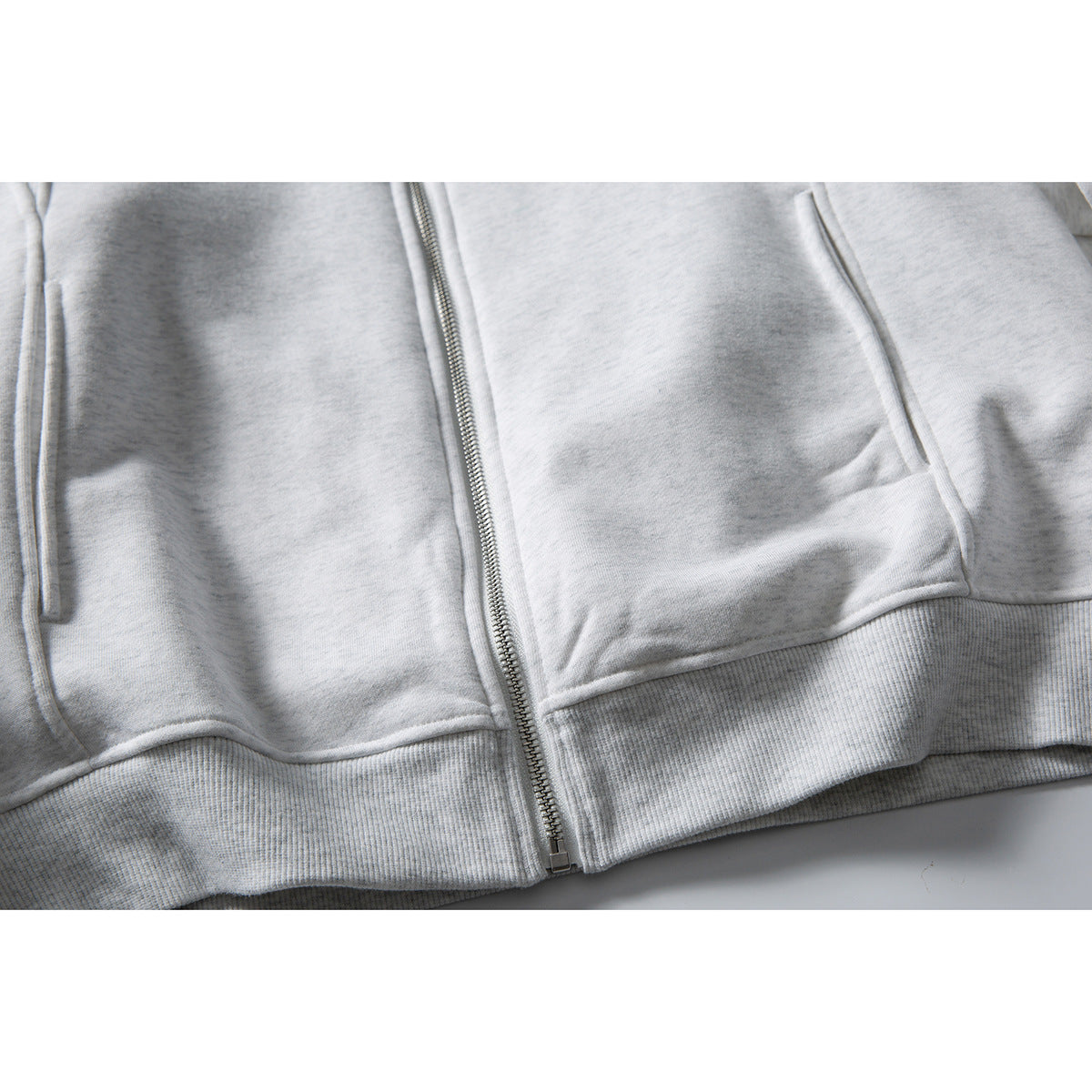 Close-up of a light gray jacket's front zipper, lower pockets, and ribbed hem.