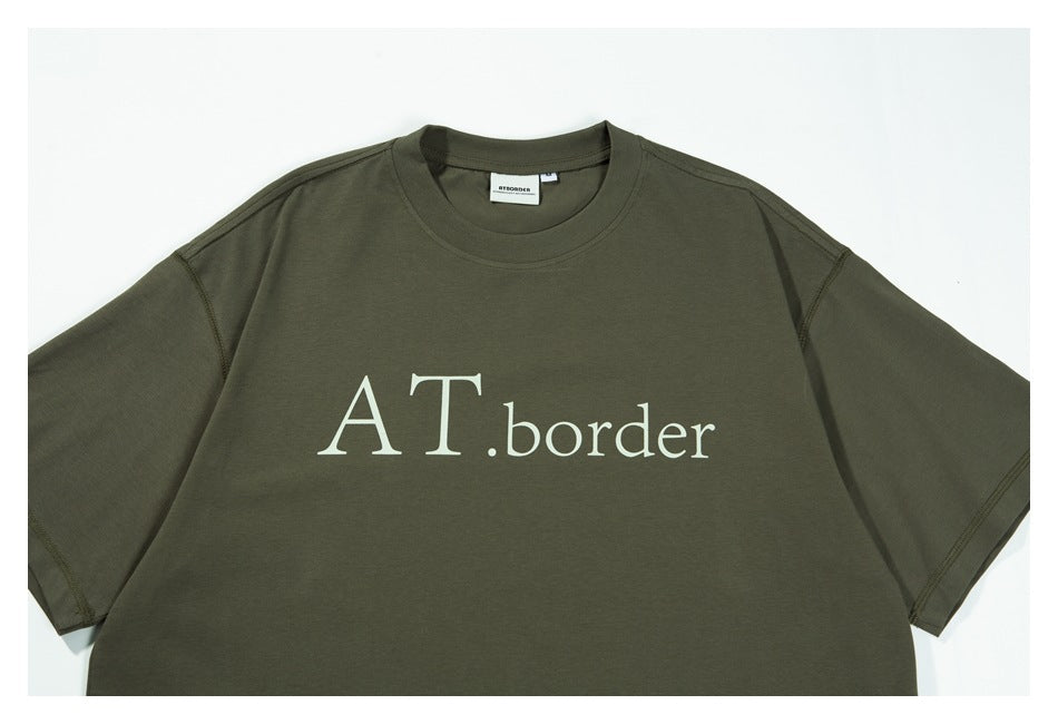 Close view of the olive-green T-shirt front with large cream “AT.border” lettering.