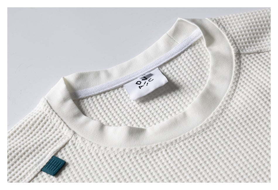 Close-up of an ivory waffle-knit top showing its ribbed crew neckline, inner label and small teal fabric tab.