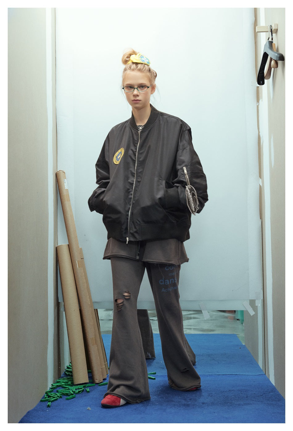 Model wearing a black zip-front bomber jacket with a round chest patch and zippered sleeve pocket.