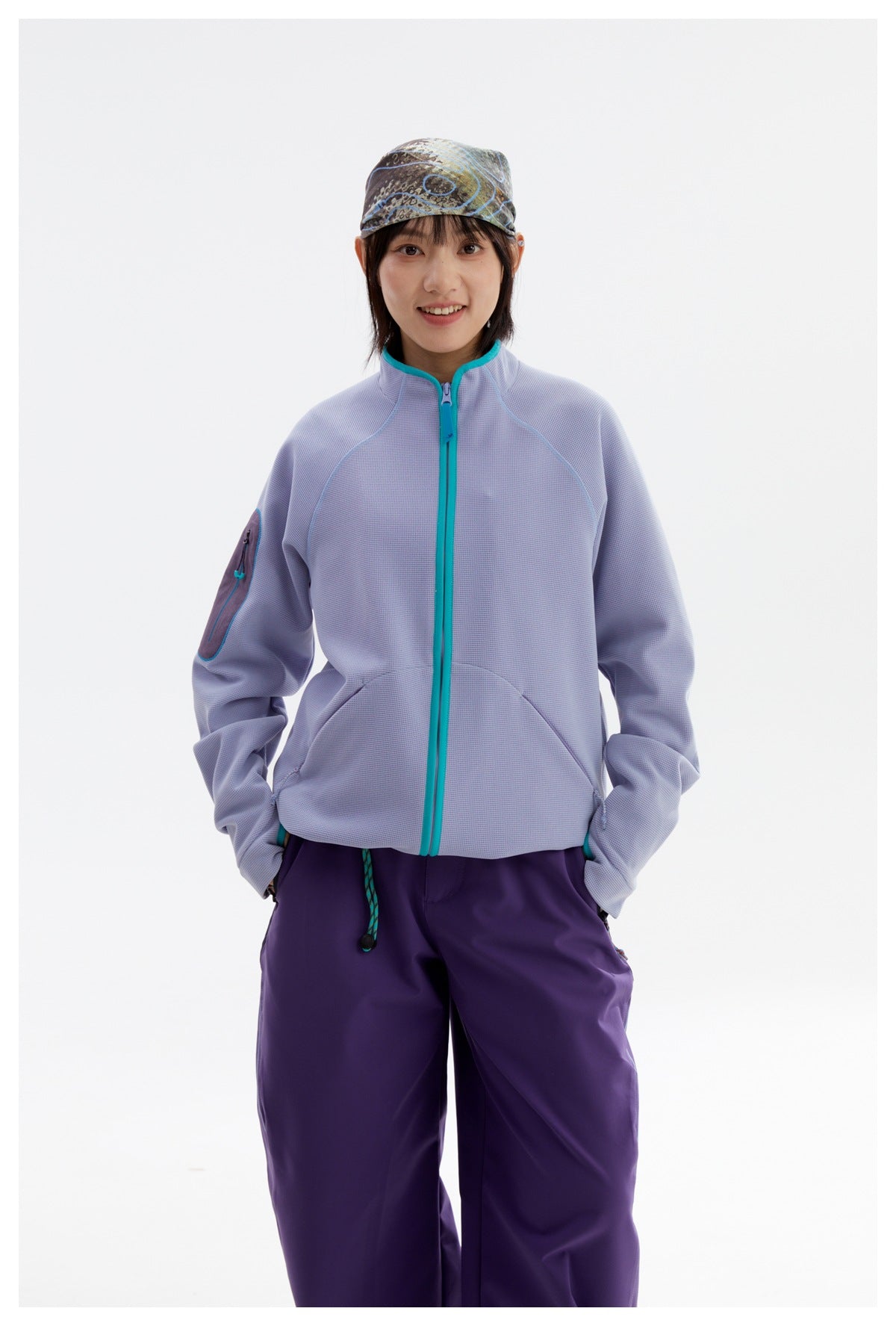 Front view of a model in a pale lavender jacket with teal zipper trim and loose purple pants.