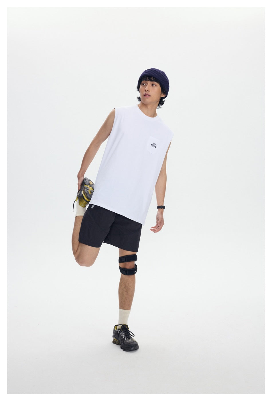 Full-length front view of a model wearing a white sleeveless pocket shirt, black shorts, and a navy beanie.