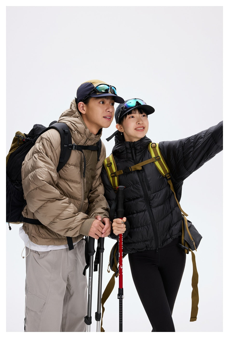 Two hikers in taupe and black hooded puffer jackets pose with backpacks and trekking poles in a close studio view.