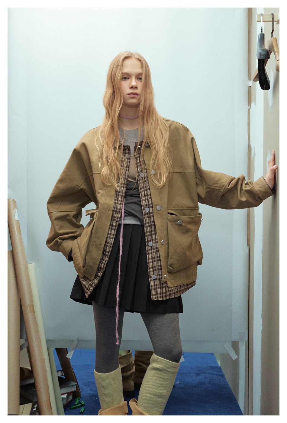 Model in an oversized olive-brown jacket with buttoned pockets and visible plaid lining, shown from the front.
