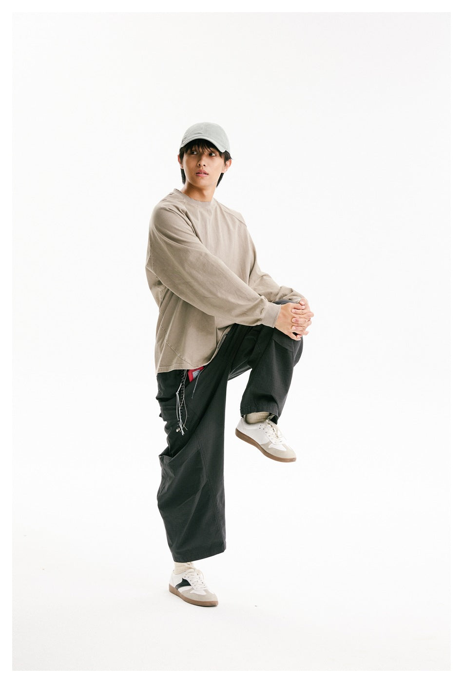 Model wearing a taupe oversized raglan sweatshirt, light cap, and dark cargo pants, posing with one knee raised.