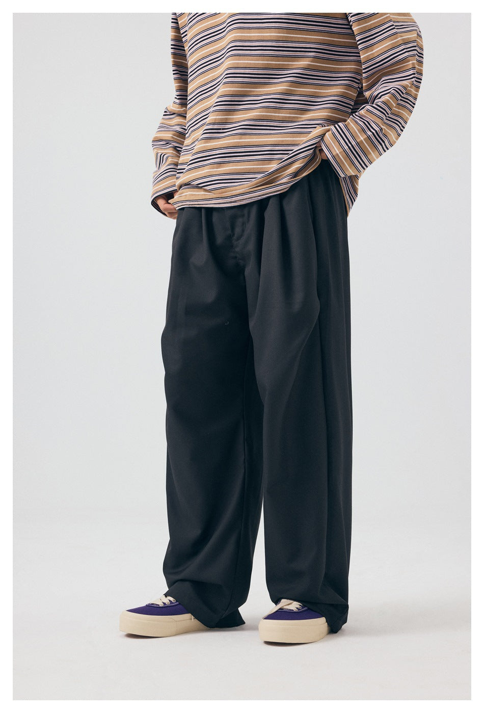 Front view of a model wearing loose black trousers with a tan striped long-sleeve top and purple sneakers.