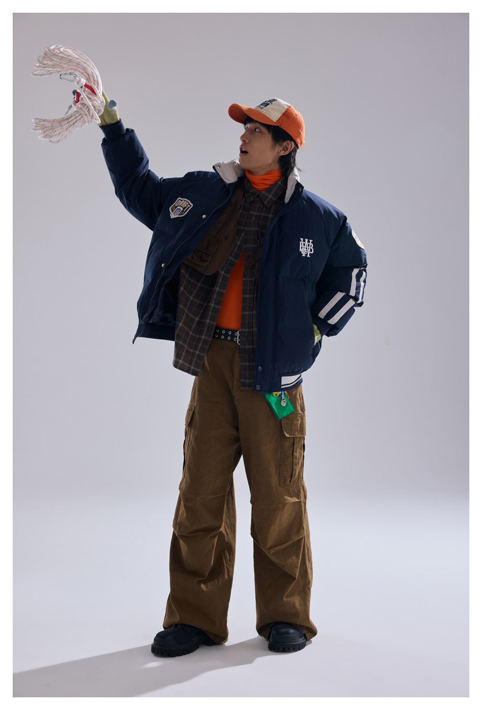 Model in navy puffer jacket with raised arm holding a rope, over plaid shirt, with brown cargo pants and orange cap