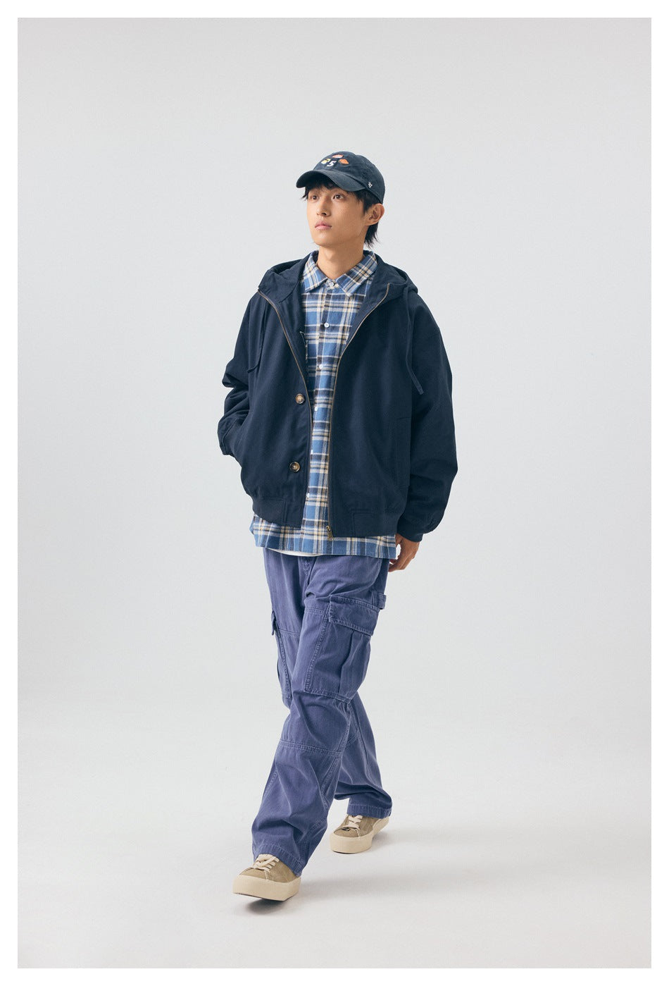 Model wearing a navy hooded zip jacket over a blue plaid shirt, with purple cargo pants and a cap.