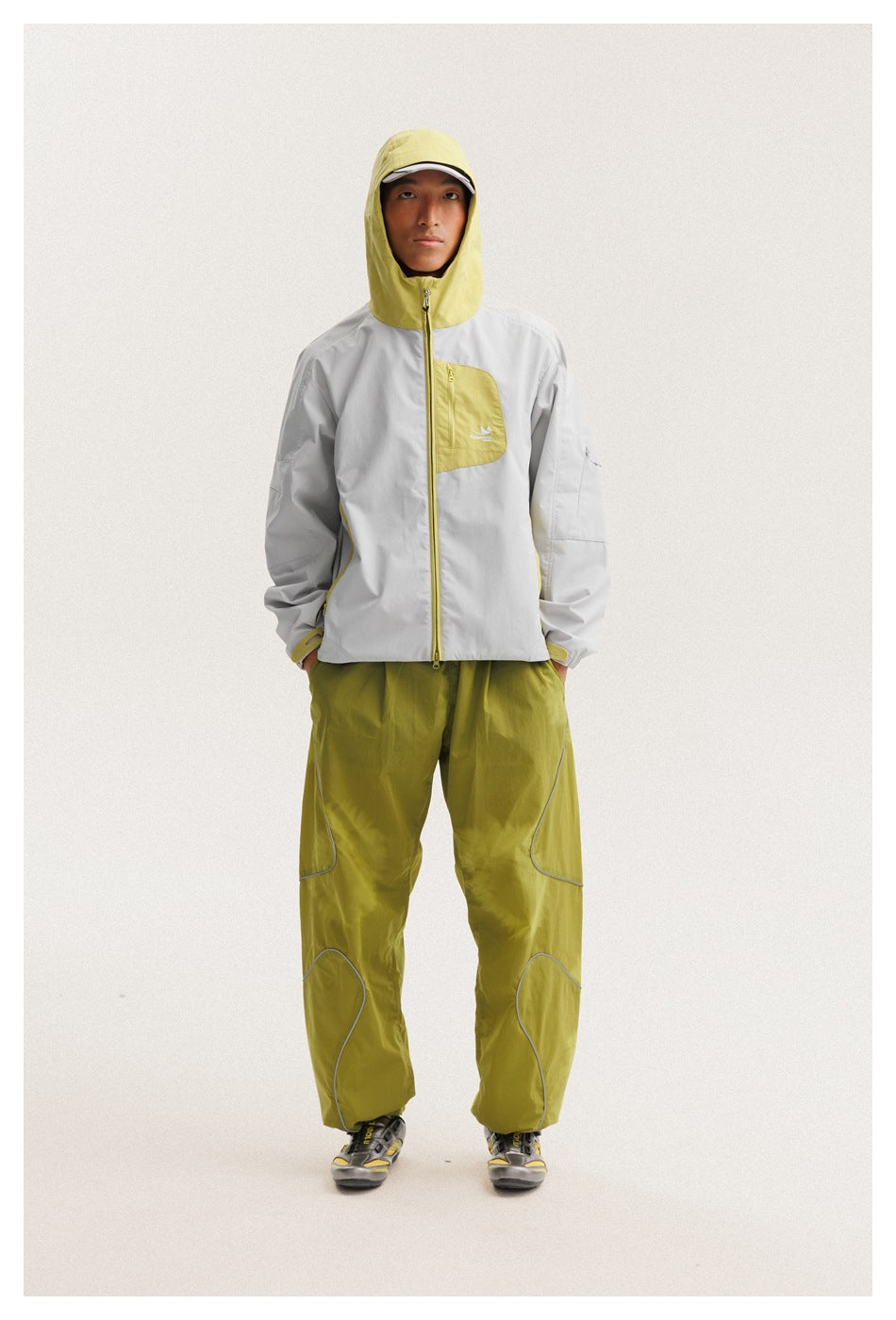 Full-length front view of a model wearing a light gray jacket with the yellow-green hood up and olive trousers.