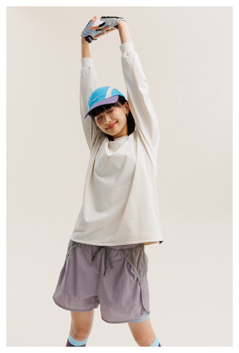 Model wearing an oversized cream long-sleeve shirt, gray-mauve shorts, a blue cap, and gloves.