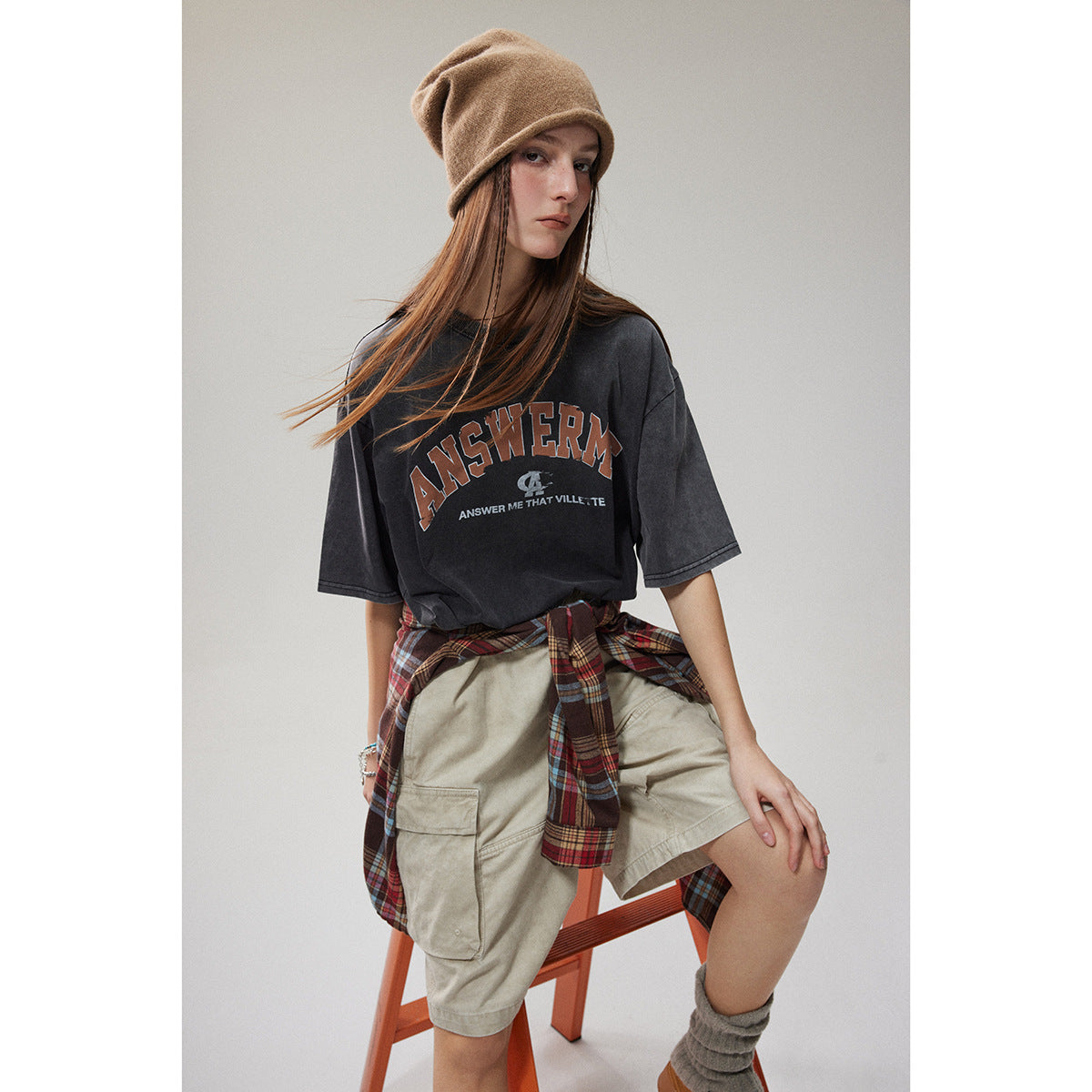 Model wearing an oversized charcoal graphic T-shirt with beige cargo shorts and a red plaid shirt tied at the waist.