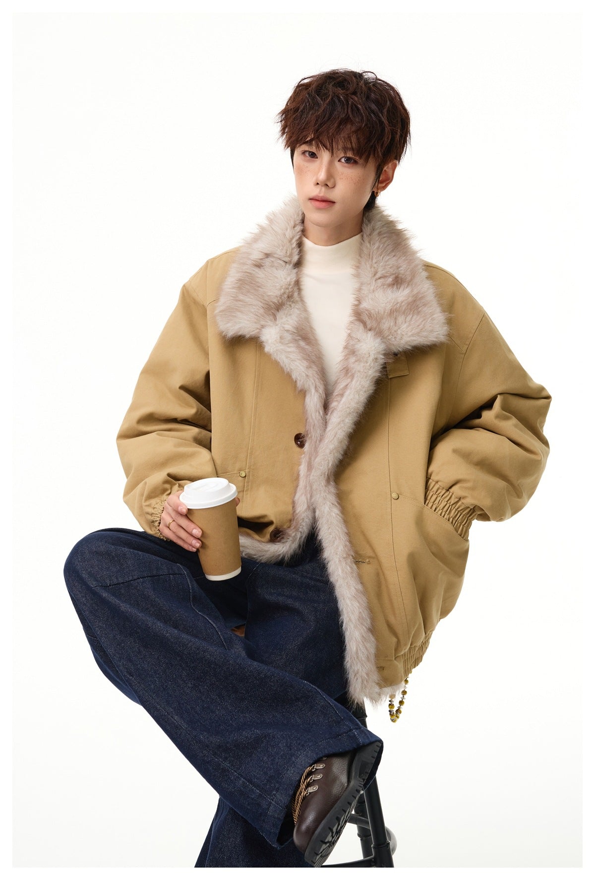 Seated model wearing a tan jacket with a wide pale fur collar and front trim, styled with dark jeans.