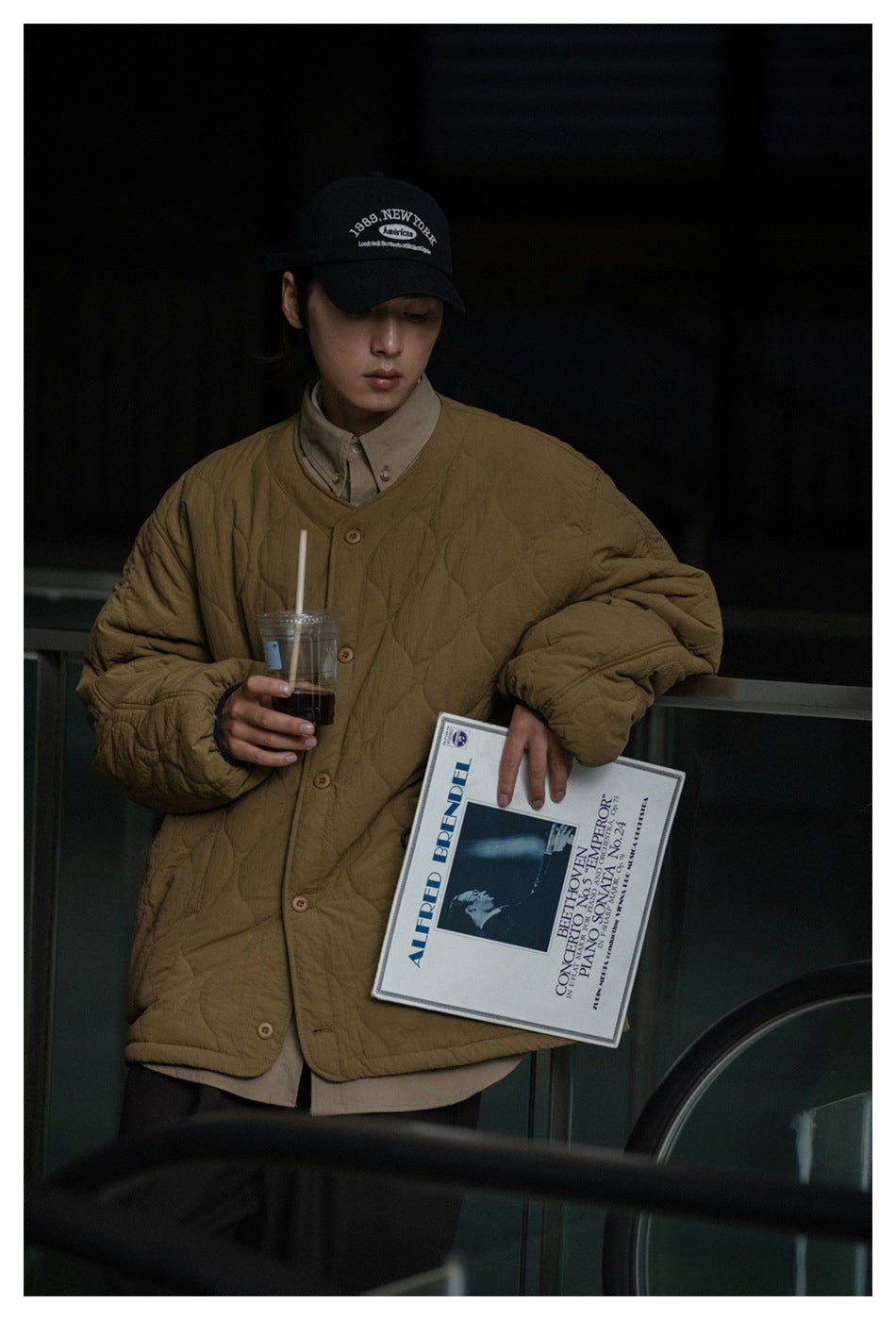 Model wearing a brown quilted button-front jacket over a beige shirt; cap and printed booklet are also visible.