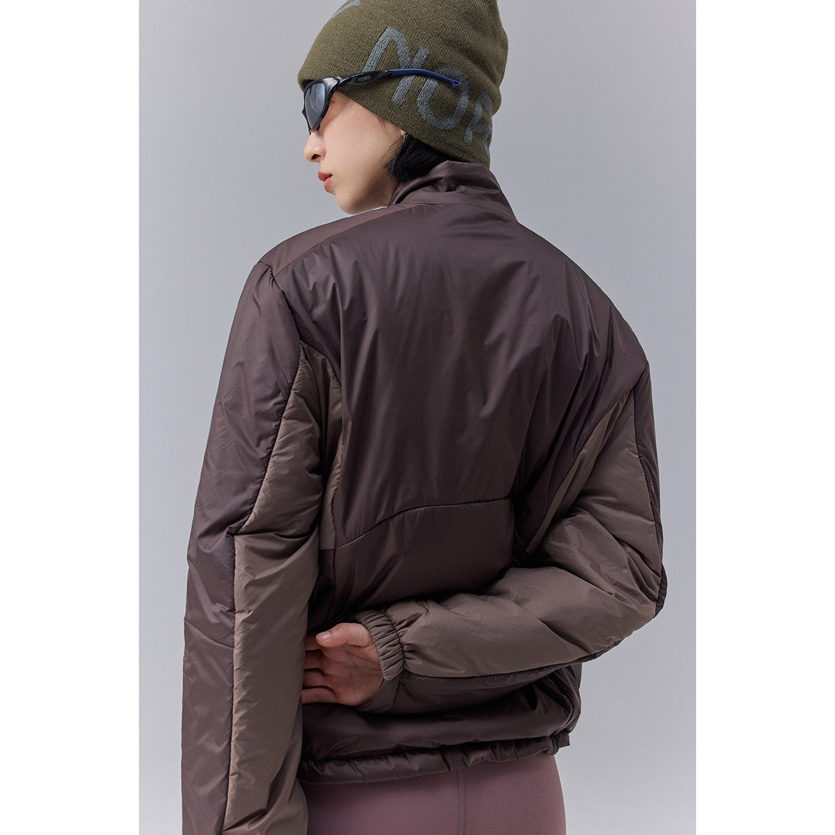 Close rear view of a dark brown quilted jacket worn by a model, showing its paneling, sleeves, and elastic cuffs.