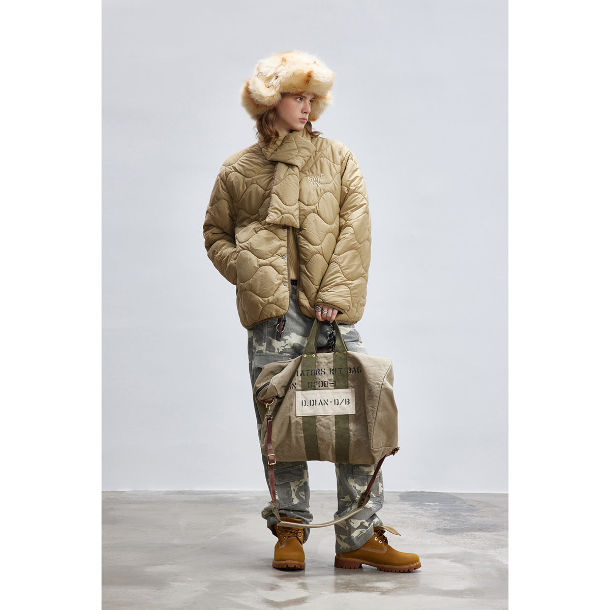 Full-length model wearing a tan quilted jacket with a padded wrap collar, camouflage trousers and a fur hat.