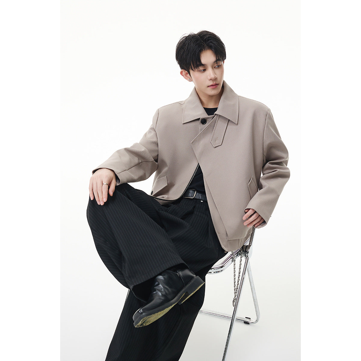 Seated model wearing a taupe jacket with a broad collar, asymmetric front and angled pockets.