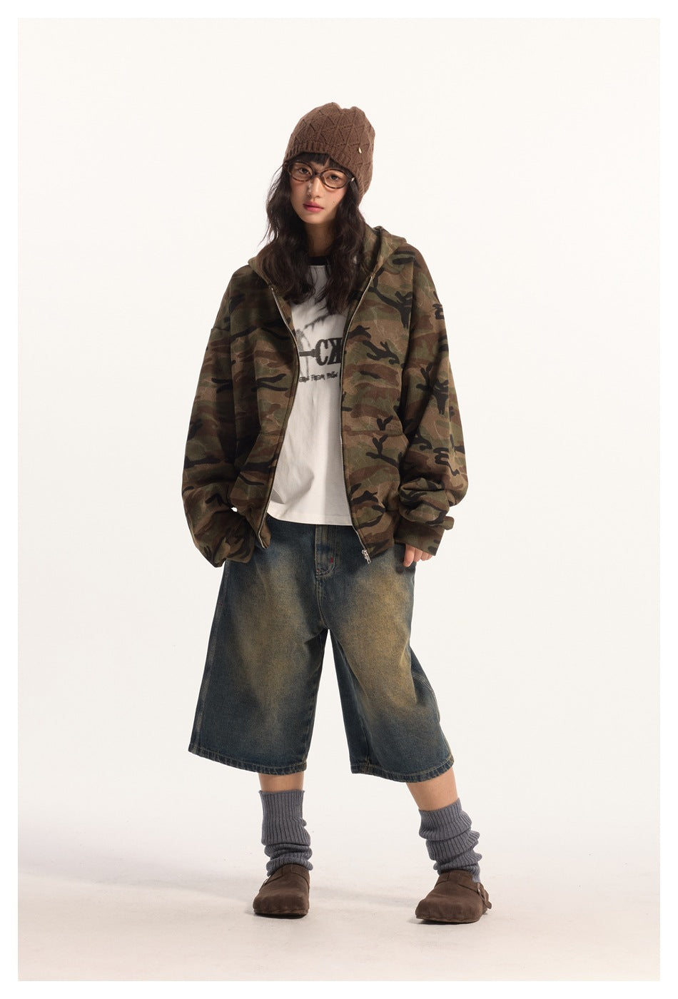 Full-length model wearing an open camouflage zip hoodie, white graphic tee and loose dark denim shorts.