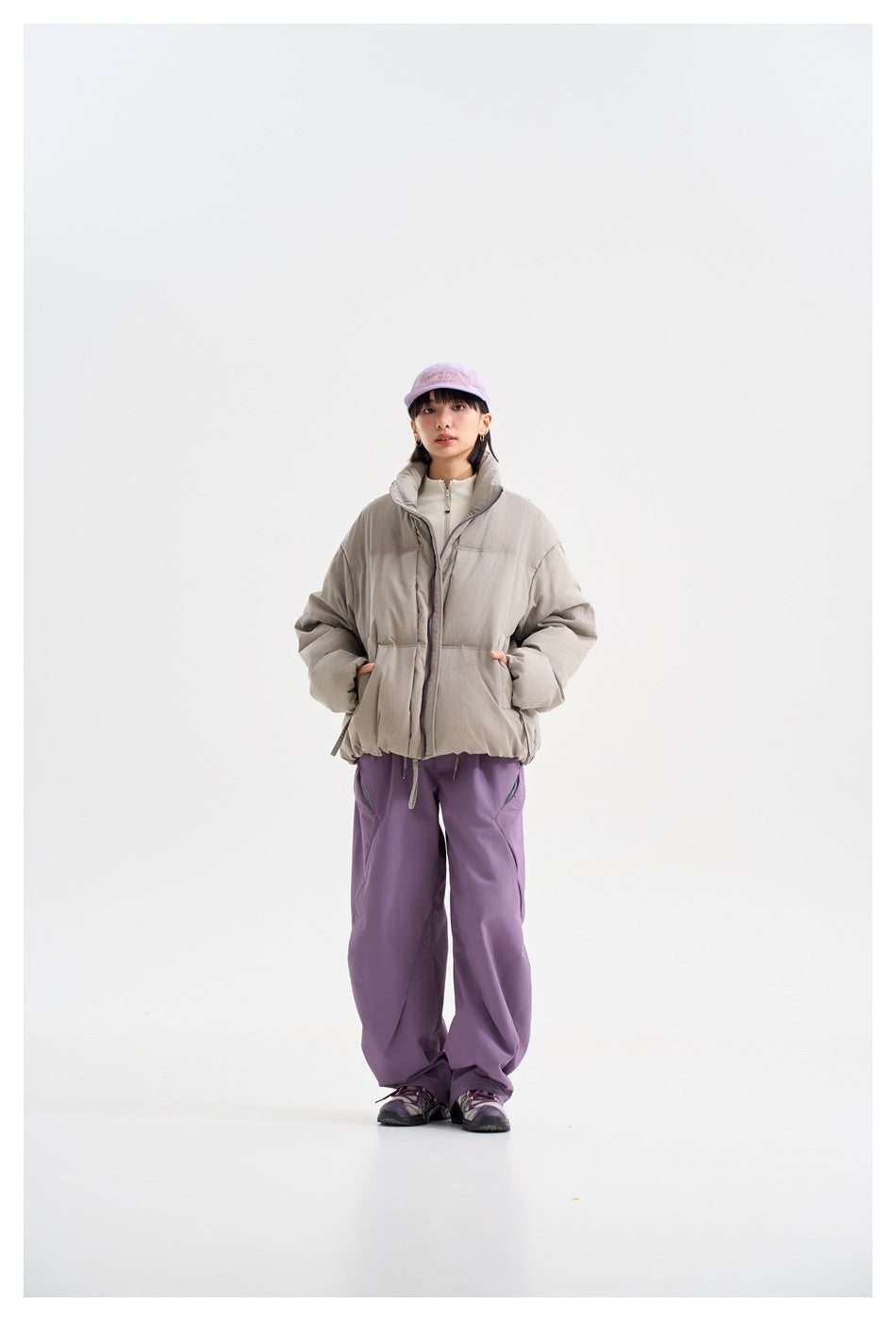 Front view of a model wearing a light gray puffer jacket with purple trousers and a lavender cap.