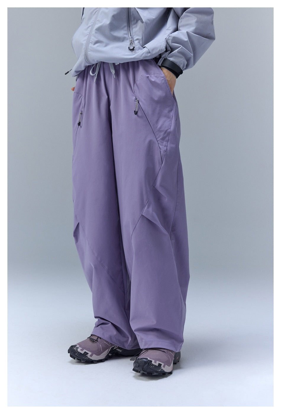 Front view of a person wearing loose lavender pants with drawstring details and zip pockets.