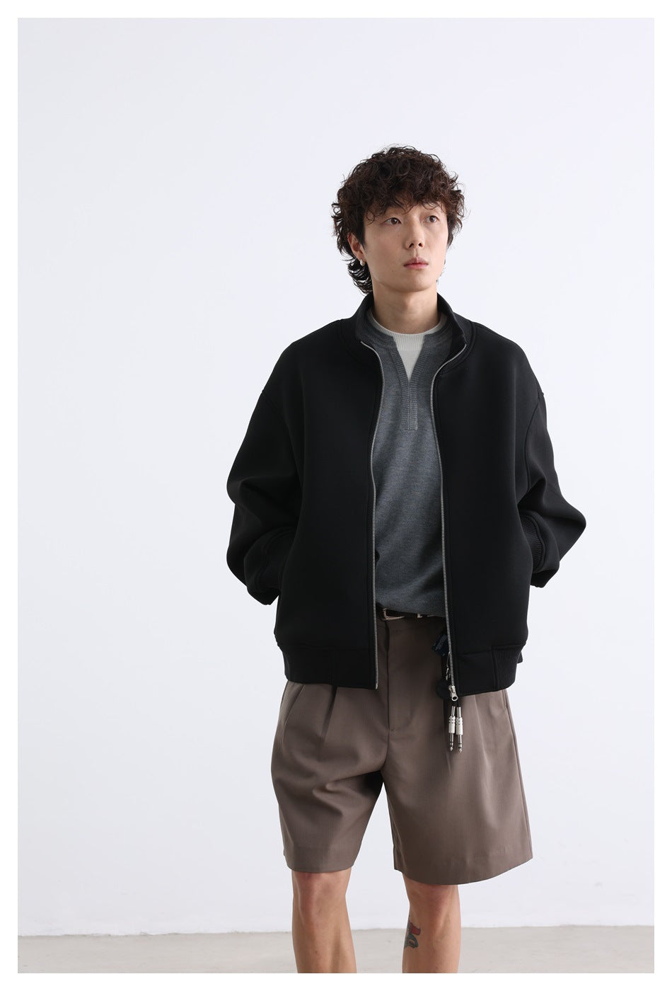 Model wearing an open black zip-front jacket with a stand collar and taupe shorts, shown from the front.