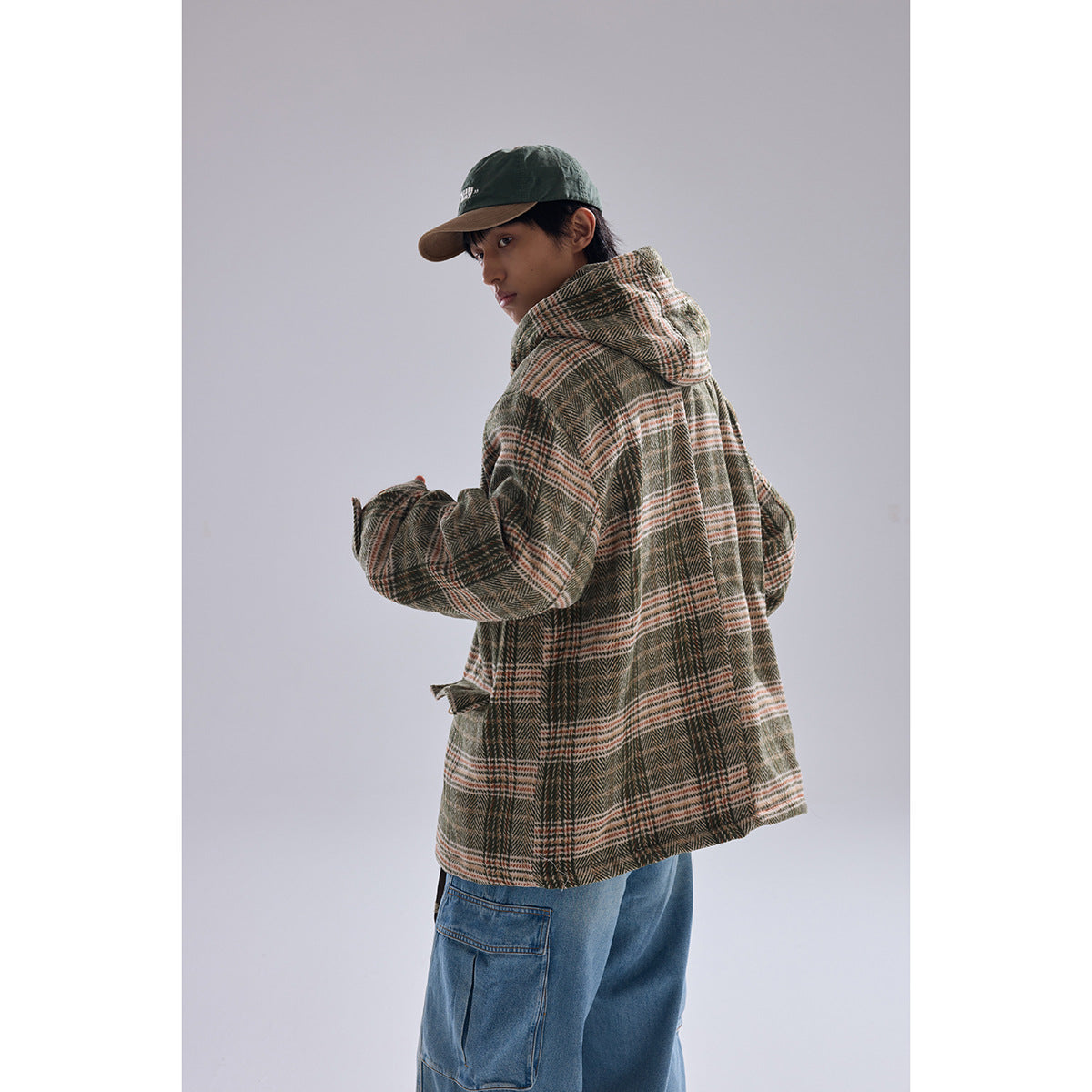 Model wearing a brown, olive, and cream plaid hooded jacket with toggle closures and large patch pockets.