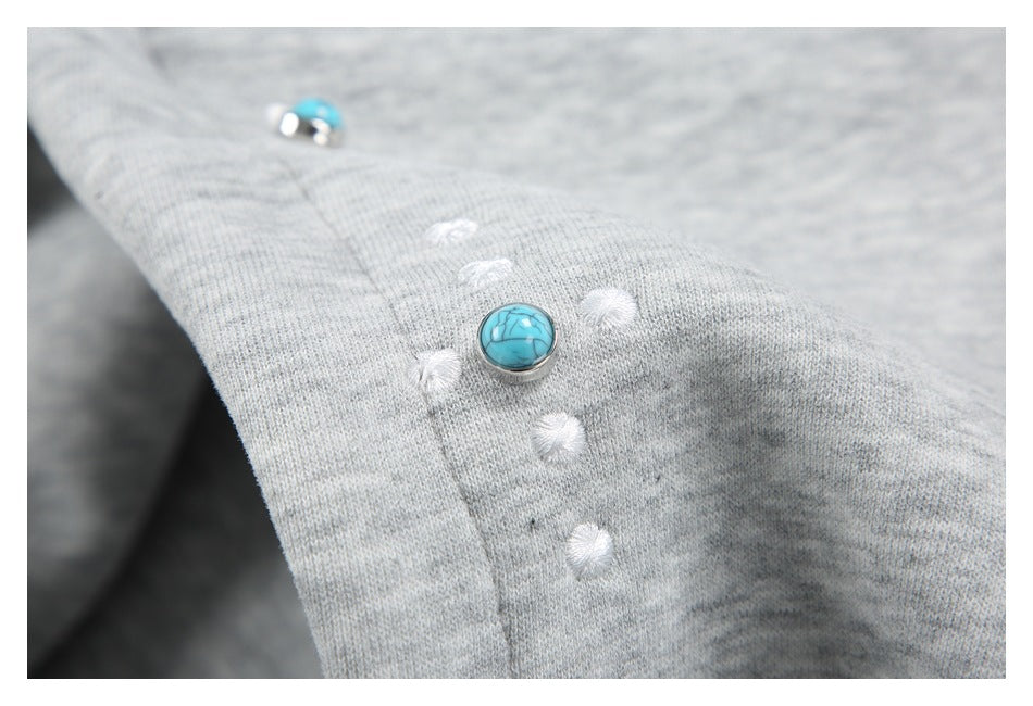 Close-up of a turquoise stud and white embroidered dots on light gray hoodie fabric.