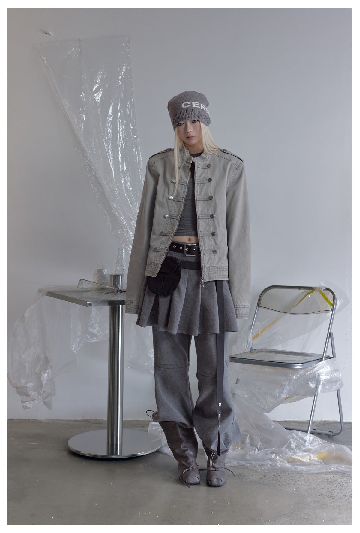 Model wearing open gray military-style zip jacket over a gray top, pleated skirt, pants and beanie, standing full-length