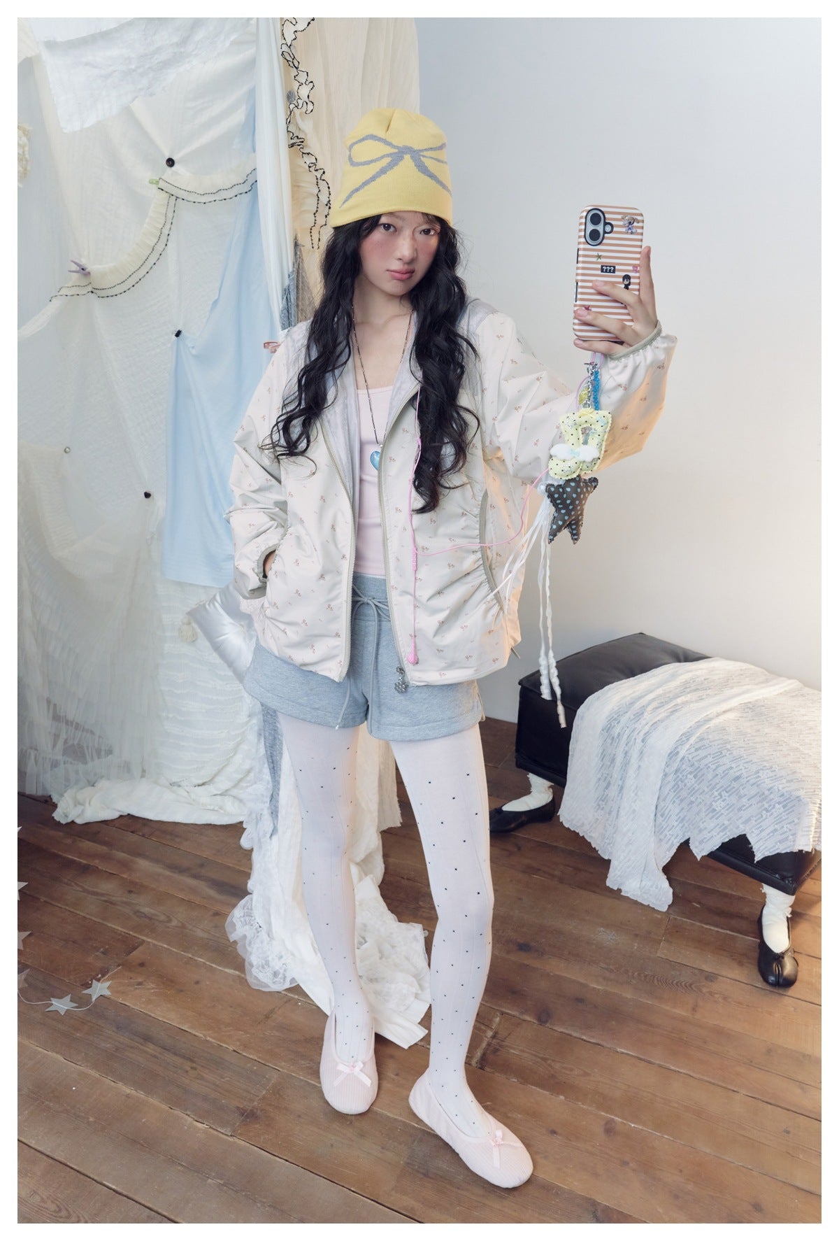 Model wearing a pale floral zip jacket, pink top, gray shorts, white dotted tights and a yellow bow-pattern beanie.