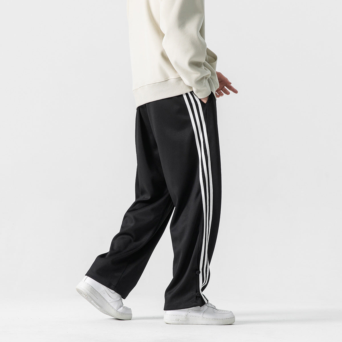 Rear three-quarter view of a model wearing black track pants with three white side stripes and snap hems.