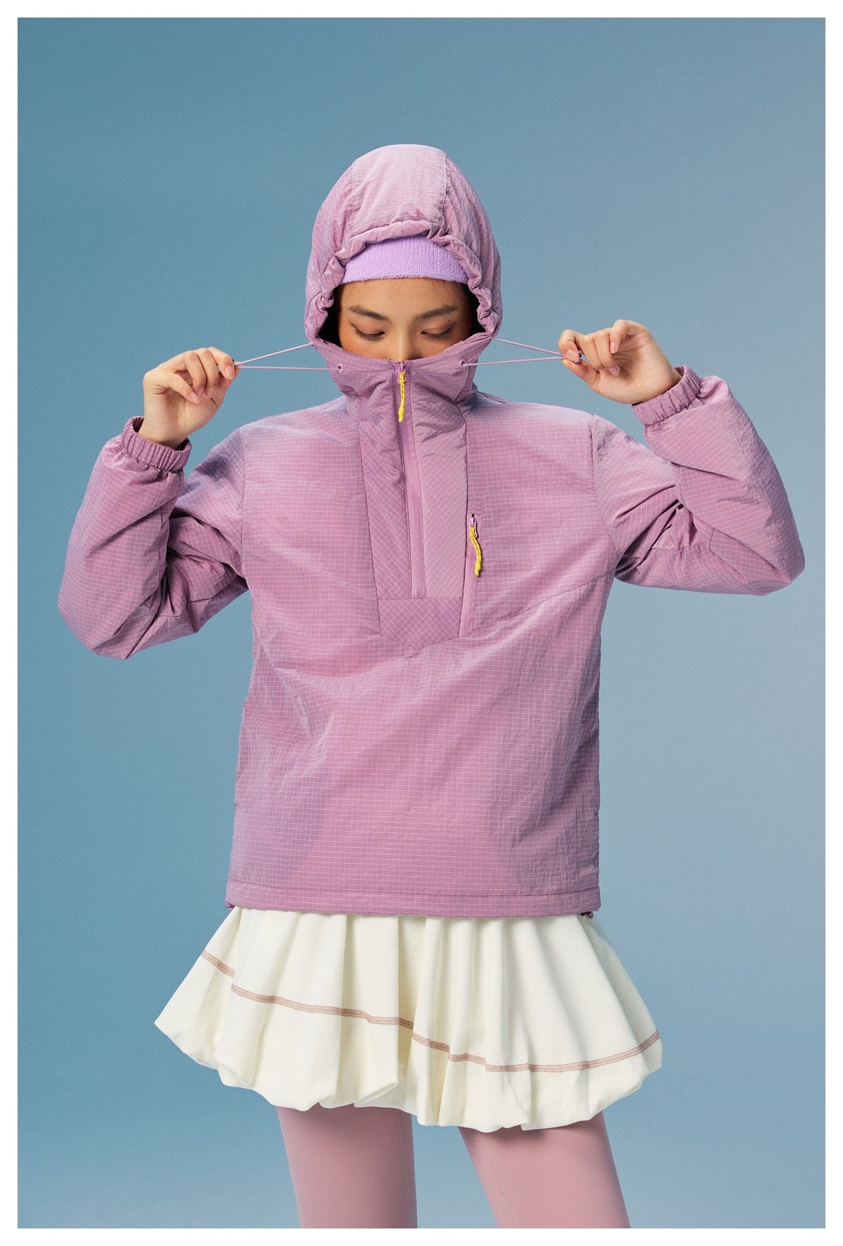 Model adjusts the hood drawcords of a lilac grid-pattern pullover with yellow zipper pulls.