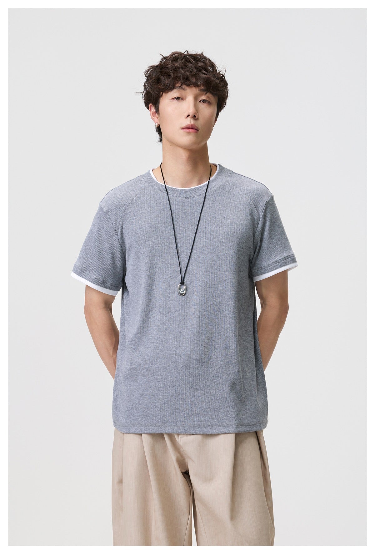 Man wearing gray layered-look short-sleeve T-shirt with pendant necklace and beige trousers, front view