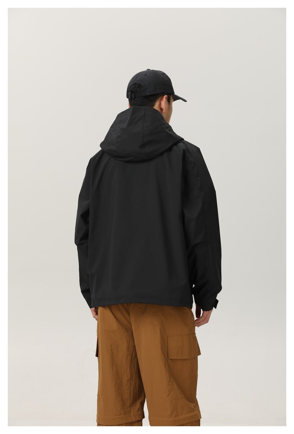 Back view of a model in a black hooded jacket and brown cargo trousers, shown within a white border.