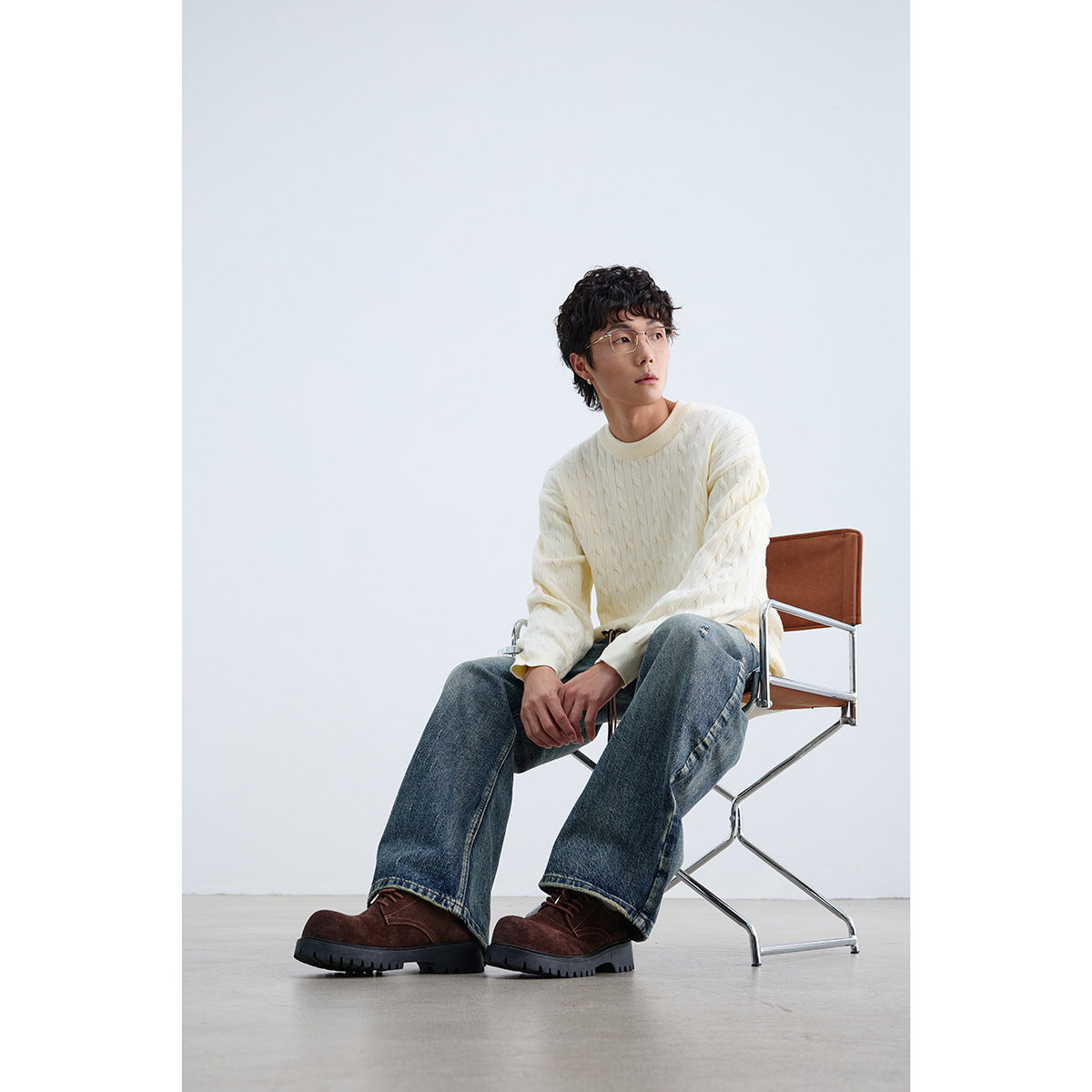 Seated model wearing an ivory cable-knit crewneck sweater and loose blue jeans.