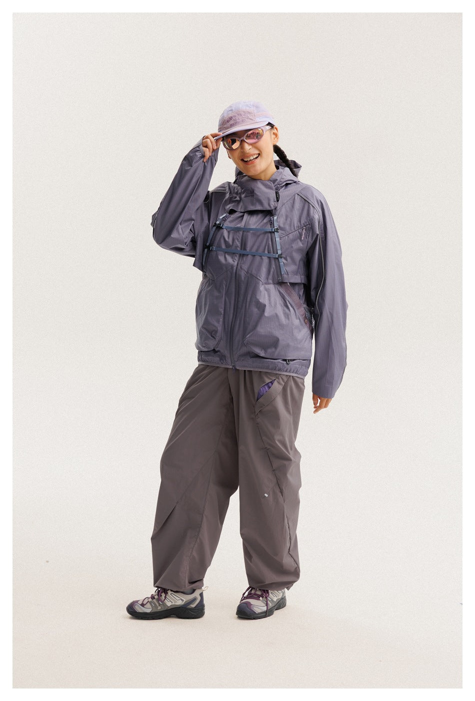 Model in a lavender-gray hooded jacket with chest straps and loose dark-gray pants, tipping the brim of her cap.