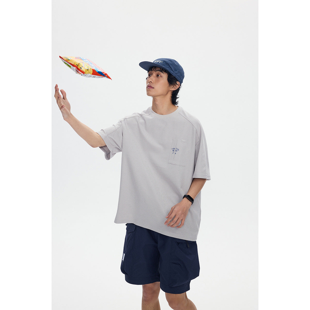 Model in a light gray oversized pocket T-shirt and navy shorts, tossing a snack packet.