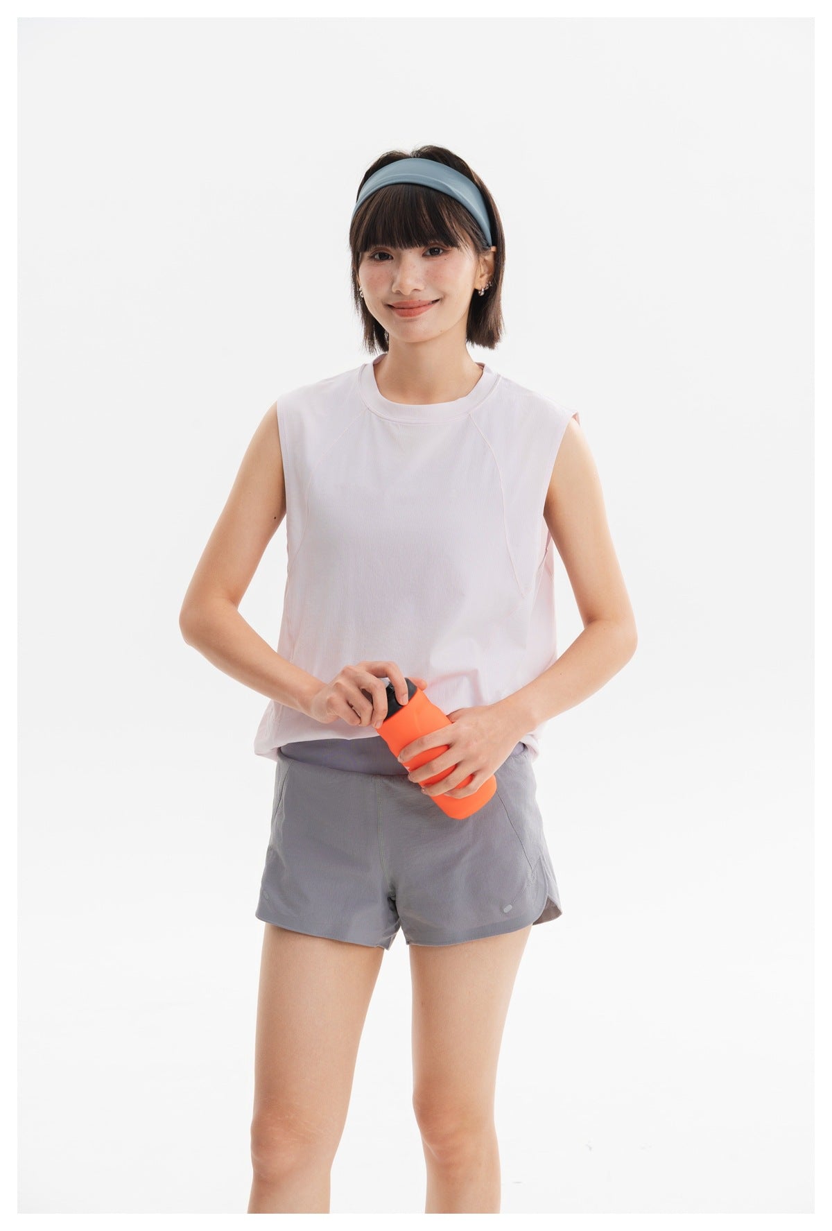 Model wearing a pale pink sleeveless athletic top and gray running shorts, holding an orange bottle.