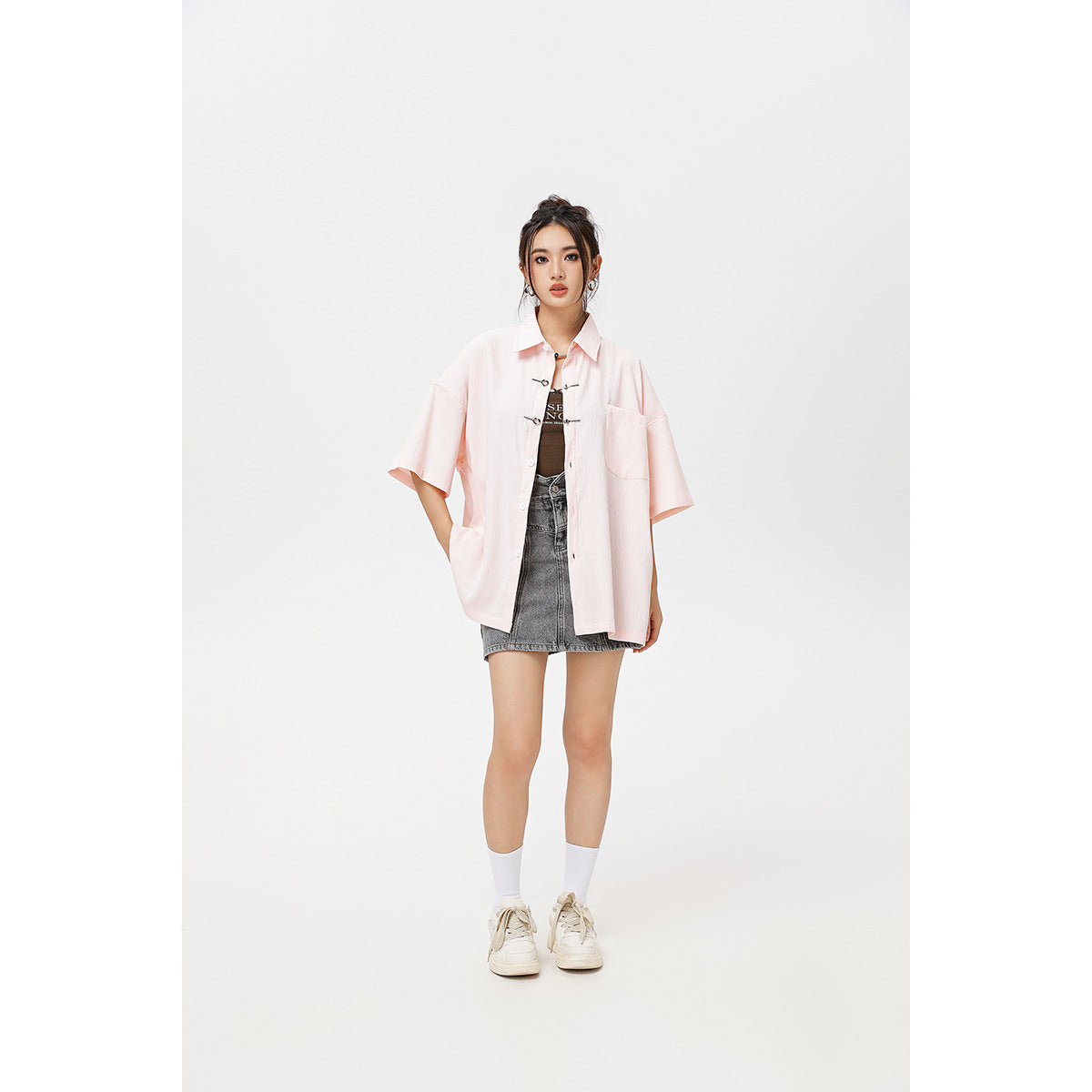 Model wearing a pale pink oversized short-sleeve shirt with metal toggles over a dark top and gray denim mini skirt.
