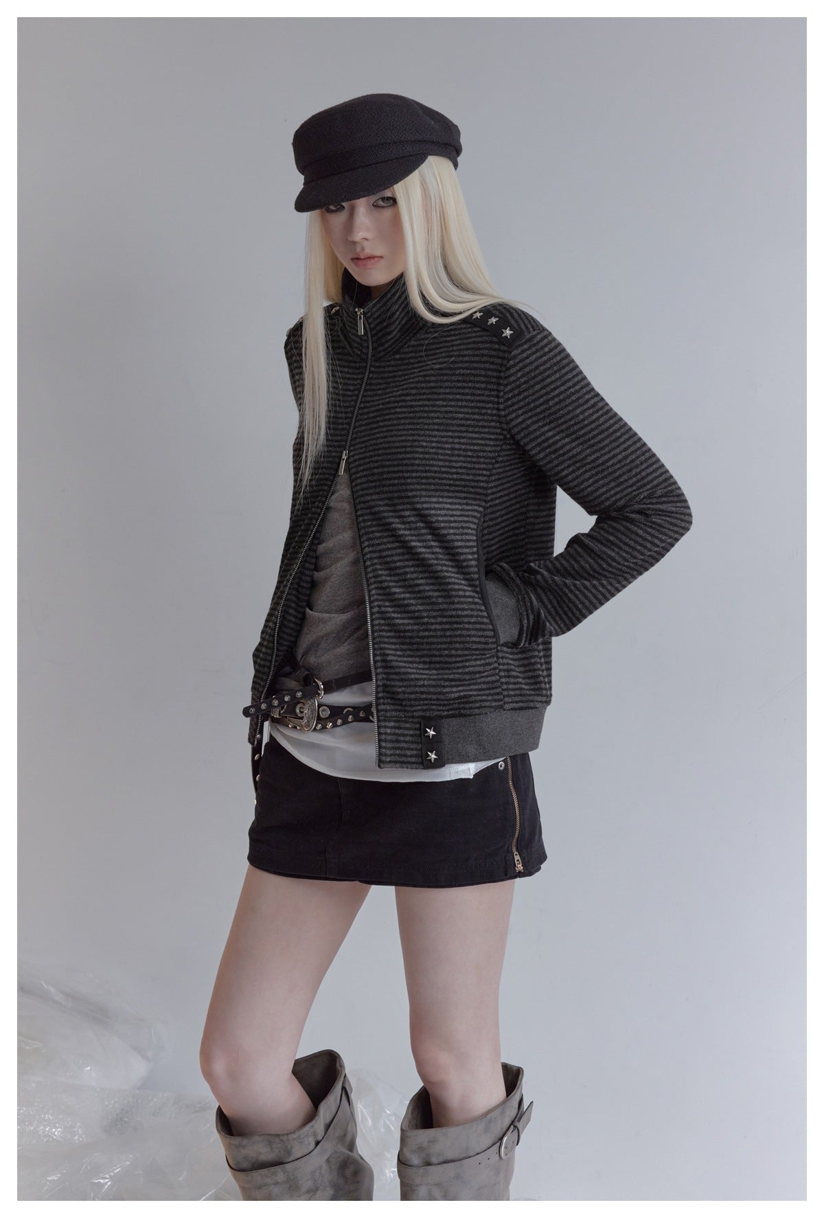 Model wearing a charcoal striped zip-up jacket with silver star studs, styled with a black cap and skirt.