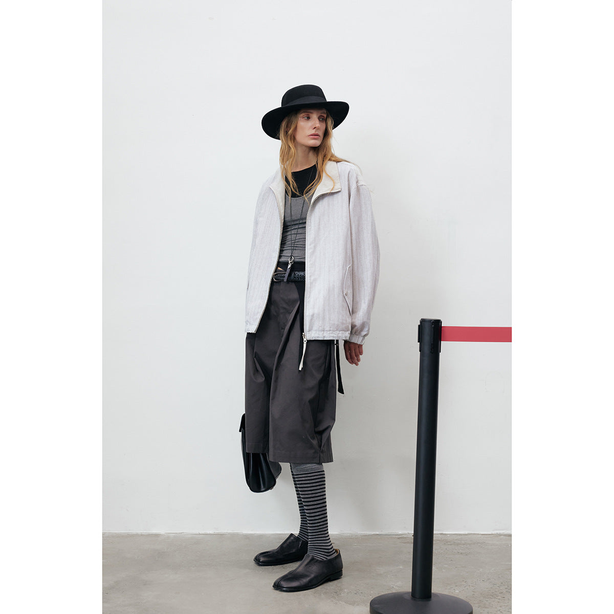Model wearing a light gray pinstriped zip jacket with dark wide-leg shorts, striped socks and a black hat.