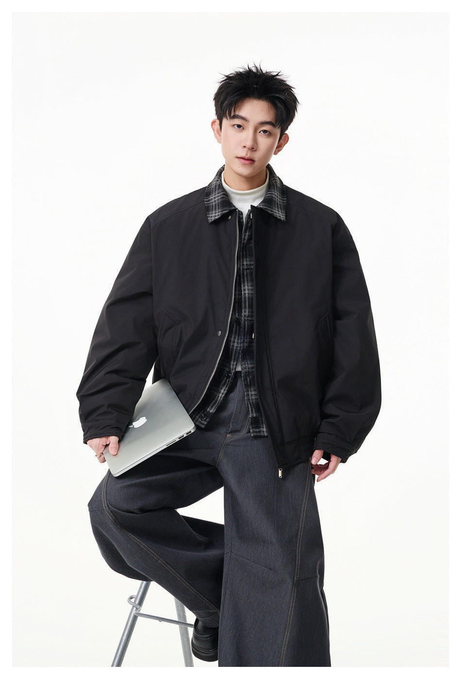Model wearing a black zip-front jacket with a plaid collar over a plaid shirt, paired with wide dark jeans.