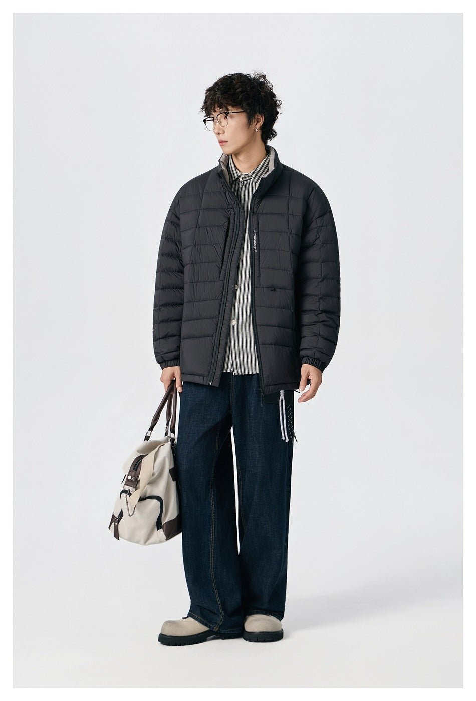 Model wearing a black quilted zip jacket over a striped shirt, with dark wide-leg jeans and a light canvas bag.