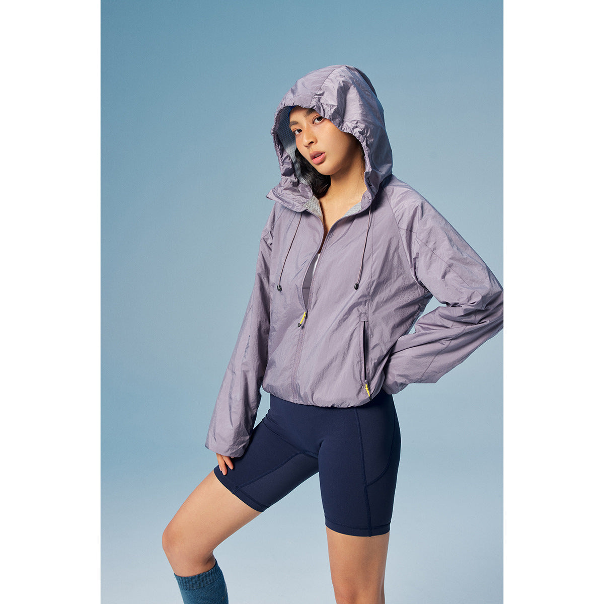 Model wearing a lavender hooded windbreaker with drawstrings and zip pockets, paired with navy cycling shorts.