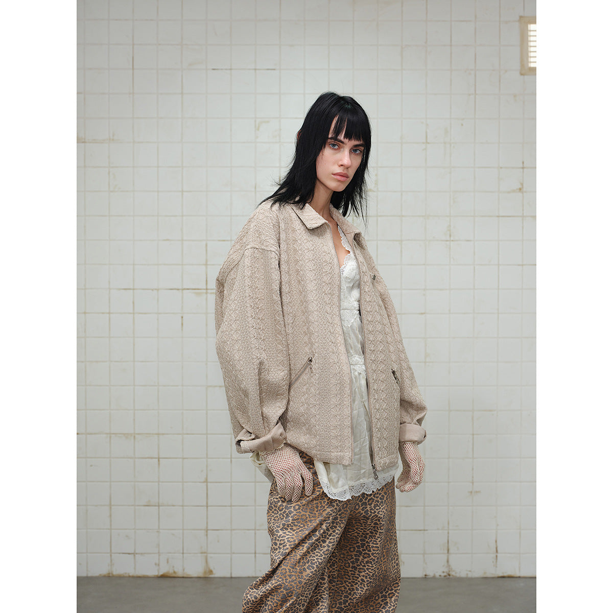 Closer angled view of a model in a light beige textured zip jacket, white lace top, leopard-print pants and patterned gloves.