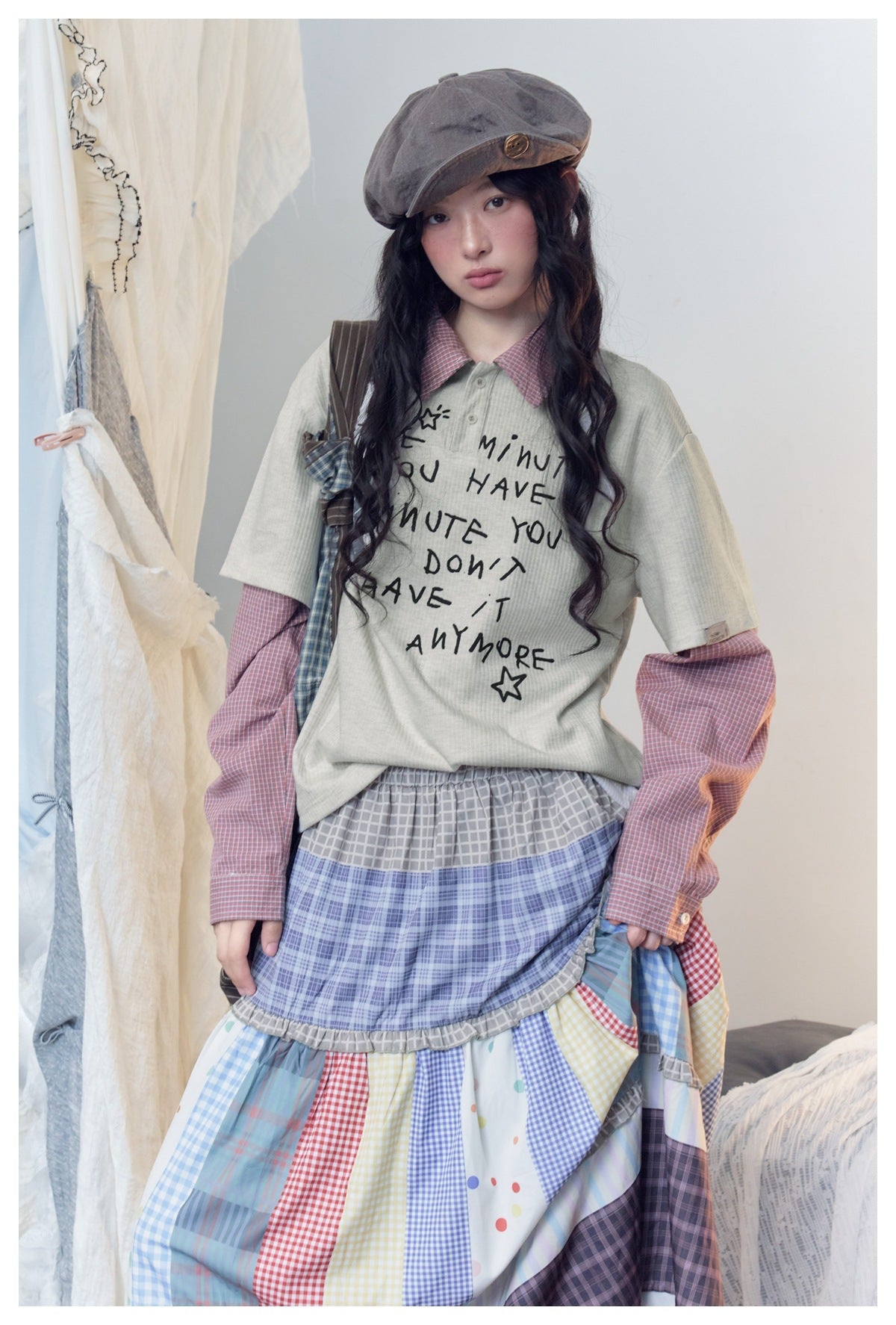 Model wearing a pale gray graphic tee over a pink checked shirt and a long tiered multicolor plaid skirt.